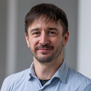 Mann mit kurzem Bart und braunen Haaren, die leicht auf die Stirn fallen sowie hellblauen Hemd lächelt zur Kamera. 