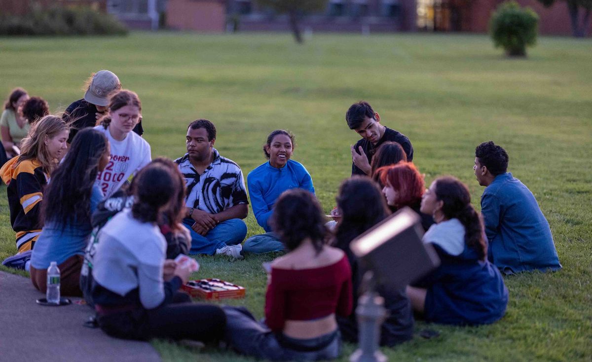 A group of students is sitting in a circle on the lawn, some are playing cards.