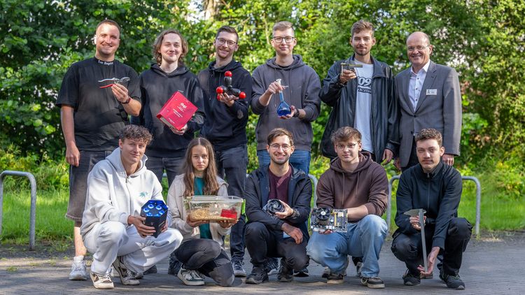 Das Bild zeigt zehn der 15 Auszubildenden. Sie stehen vor einem Unigebäude, im Hintergrund befinden sich grüne Büsche. Meik Möllers steht rechts im Bild. Alle lächeln in die Kamera. Die Azubis halten verschiedene Gegenstände in ihren Händen, zum Beispiel eine Kneifzange, ein Buch mit Verwaltungsvorschriften, eine Lampe oder einen Laborkolben.