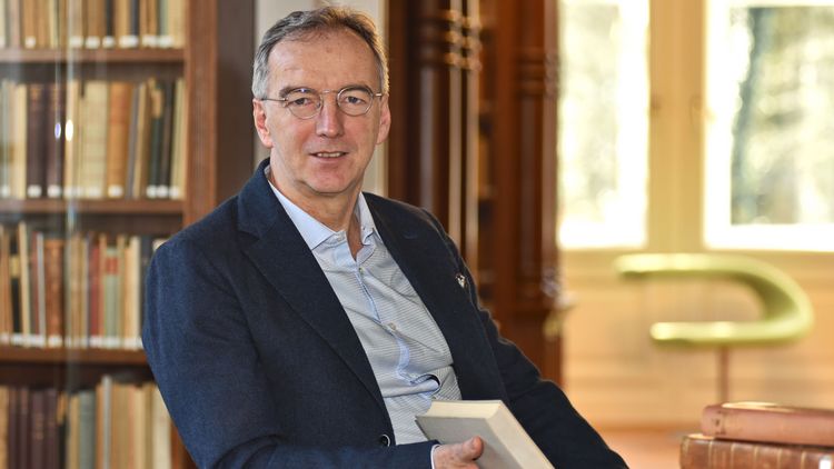 The picture shows Matthias Bormuth. He is sitting in front of a bookshelf in the Karl Jaspers House with a book in his hand and smiling at the camera.