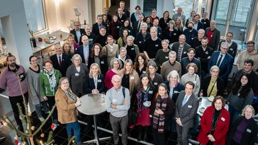 Das Bild zeigt die Mitglieder der Senate und Uni-Leitungen beider Universitäten auf einem großen Gruppenbild. Die Kamera steht leicht über ihnen, sodass alle Leute leicht nach oben schauen. Im Hintergrund sieht man ein Buffet.