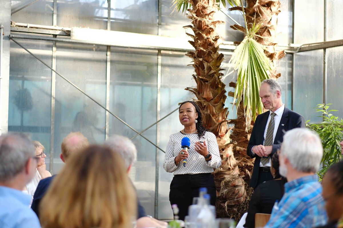 Das Bild zeigt eine Frau, die vor einer Gruppe von Personen spricht. Sie hält ein Mikrofon in der Hand und scheint eine Rede zu halten. Neben ihr steht ein Mann im Anzug und hört aufmerksam zu. Die Szene spielt in einem hellen Raum mit großen Fenstern und tropischen Pflanzen im Hintergrund auf dem Gelände des Botanischen Gartens. Es handelt sich um das Welcome Dinner im Rahmen des 25-jährigen Kooperationsjubiläums der Universität Oldenburg mit der Nelson Mandela University. 