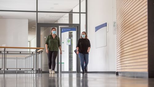 Zwei junge Frauen mit Maske laufen nebeneinander durch das Hörsaalgebäude A14.