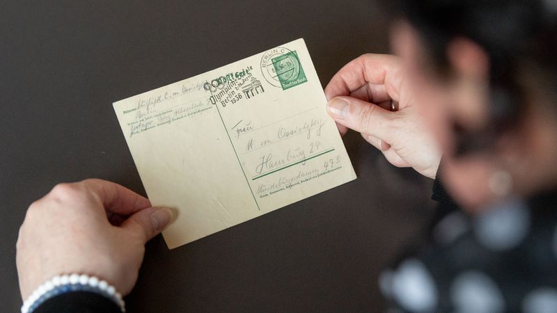 Vorderseite der Postkarte, die von zwei Händen gehalten wird. Man sieht die handgeschriebene Adresse, den Absender, eine grüne Briefmarke und einen Poststempel  Briefmarke