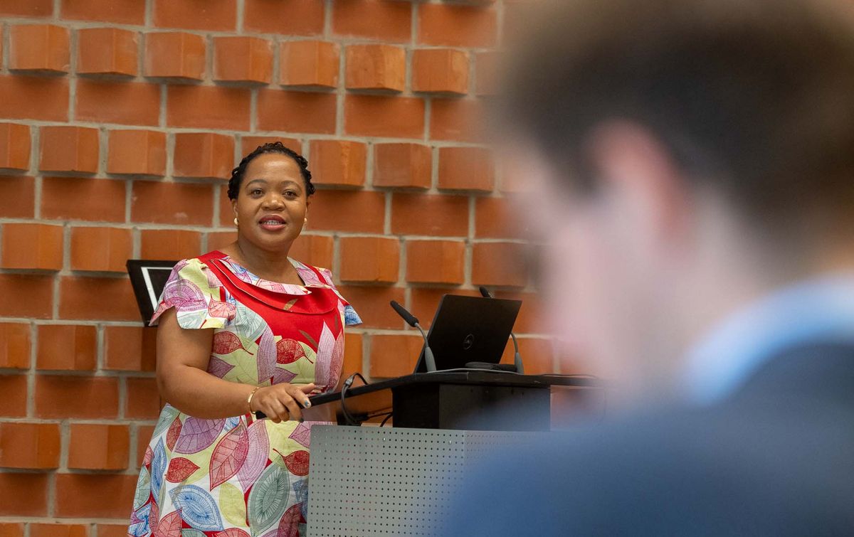 Eine Frau in einem bunt gemusterten Kleid steht hinter einem Pult und spricht. Sie lächelt und lehnt sich an das Pult. Im Hintergrund ist eine rote Backsteinwand und vor ihr eine verschwommene Person zu sehen. Das Bild vermittelt den Eindruck einer Präsentation oder Rede während des Symposiums. Die Frau scheint eine wichtige Rolle bei der Veranstaltung zu spielen und hält möglicherweise einen Vortrag.