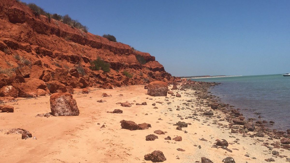 Eine Küstenlandschaft mit rotem Felsenhang, Sandstrand und türkisfarbenem Wasser.