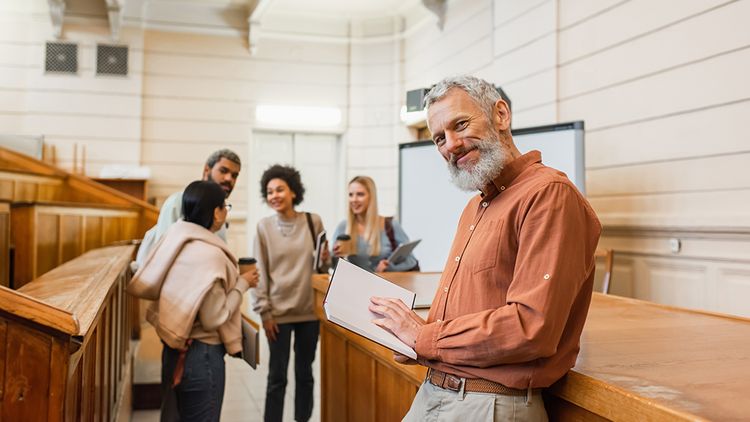 Smiling teacher holding notebook and looking at camera near blurred multiethnic students in university
