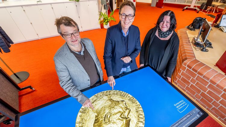 Das Bild zeigt die drei genannten Personen, die um einen Multitouch-Tisch stehen. Herr Zimmermann und Frau Andermann zeigen mit ihrem Zeigefinger auf den 3D-Scan. Alle drei Personen lächeln in die Kamera, die von schräg oben auf die Szenerie blickt.