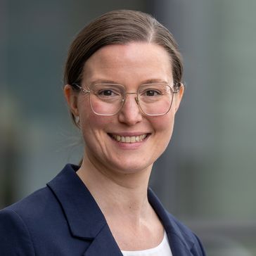 Porträt von Hannah Maier: Sie hat dunkle Haare, zusammengebunden, trägt ein weißes Shirt und einen blauen Blazer und lächelt in die Kamera.