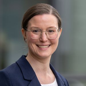 Portrait of Hannah Maier: She has dark hair, tied up, wears a white shirt and a blue blazer and smiles at the camera.