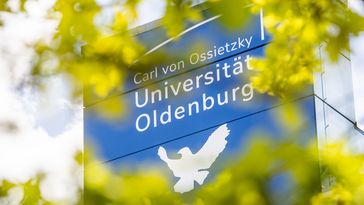 Auf dem Foto ist der blaue Turm des Gebäudes A1 bis A4 mit dem Logo der UNiversität und der Friedenstaube zu sehen, im Vordergrund sind unscharf die Blätter eines Baums zu erkennen.