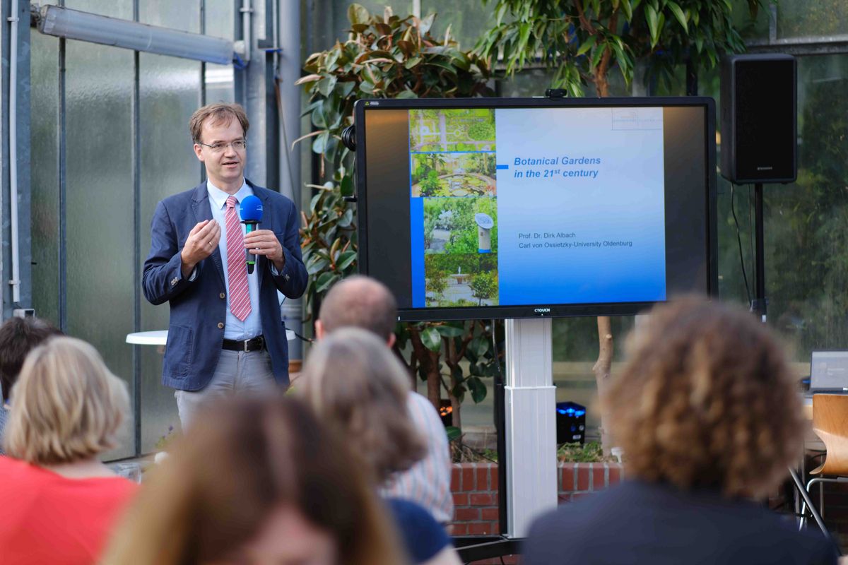 Das Bild zeigt einen Mann, der vor einer Gruppe von Personen steht und spricht. Er hält ein Mikrofon in der Hand und präsentiert eine Präsentation auf einem großen Bildschirm. Auf dem Bildschirm ist ein Bild von botanischen Gärten und der Titel "Botanical Gardens in the 21st century" zu sehen. Unter dem Titel steht der Name des Vortragenden: Prof. Dr. Dirk Albach von der Carl von Ossietzky University Oldenburg. Die Szene spielt im Rahmen des Welcome Dinners zum 25-jährigen Kooperationsjubiläum der Universität Oldenburg mit der Nelson Mandela University.