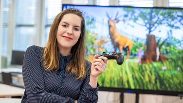 Carolin Becklas mit Gamepad vor einem Monitor auf dem ein comigartiges Spiel zu sehen ist.