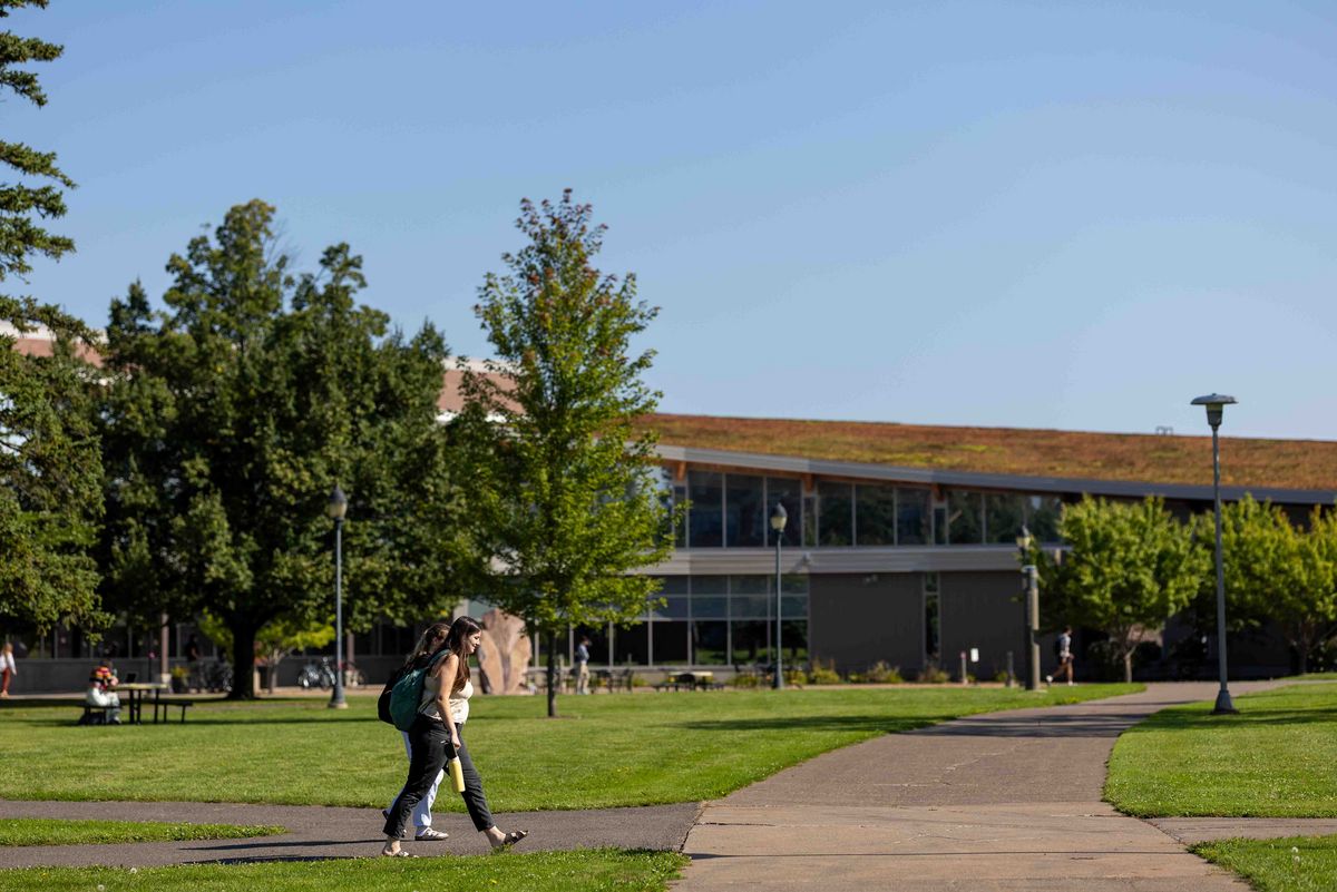 Zwei Studierende gehen auf einem Weg über den Campus. Bäume und ein Gebäude im Hintergrund.