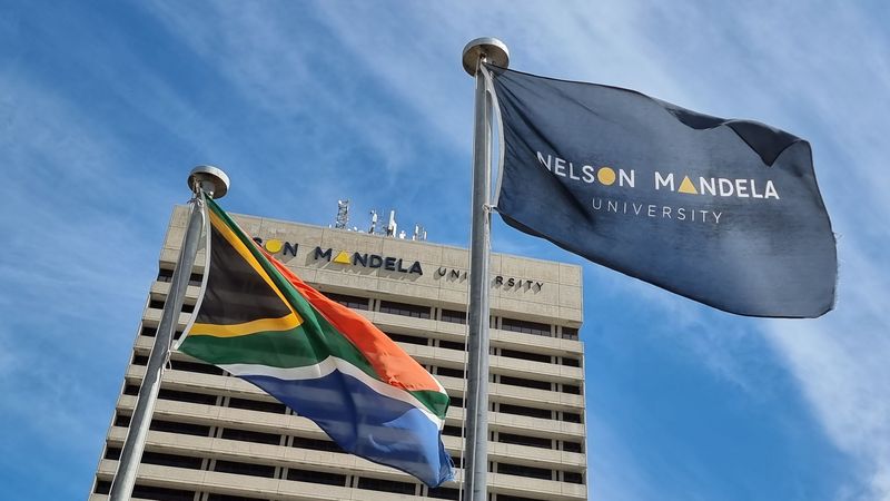 Foto der südafrikanischen Flagge und der Fahne der Nelson Mandely University (NMU) vor einem Gebäude der NMU. An den Seiten und im Hintergrund ist blauer Himmel mit leichten Schleierwolken zu erkenenen.