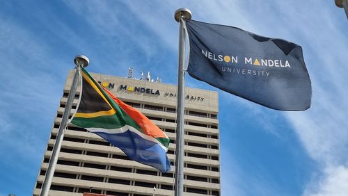 Photo of the South African flag and the flag of Nelson Mandely University (NMU) in front of a university building. The sky is blue sky with light veil clouds.