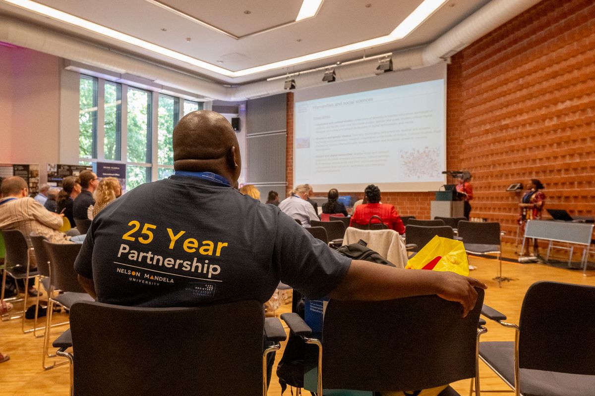 Das Bild zeigt einen Innenraum, den BIS-Saal der Universität Oldenburg. Im Vordergrund ist der Rücken einer Person zu sehen, die ein dunkelblaues T-Shirt trägt. Auf dem T-Shirt steht in großen, gelben Buchstaben "25 Year Partnership" und darunter die Logos der Nelson Mandela University und der Universität Oldenburg. Im Raum sitzen noch viele weitere Personen und blicken auf eine Person, die an einem Pult steht und eine Präsentation hält. Die Präsentationsthemen scheinen sich um Geistes- und Sozialwissenschaften zu drehen. 