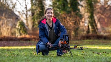 A woman is crouching in a meadow, with a drone standing in front of her.  