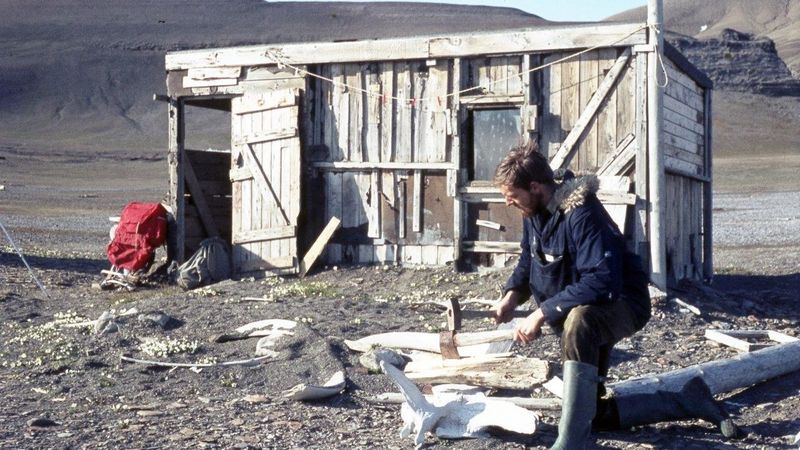 Vor einem Holzschuppen auf Edgeøya kniet Arktisforscher Paul W.J. de Groot und hackt Holz mit einem Walwirbel als Hackklotz. Links am Schuppen lehnt ein roter Rucksack, im Hintergrund sind kahle Berghänge der Insel zu erkennen.