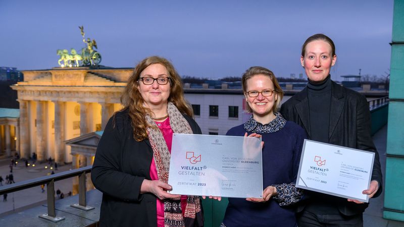 Die drei Frauen stehen auf einem Balkon mit dem Zertifikat in den Händen, im Hintergrund das Brandenburger Tor. 