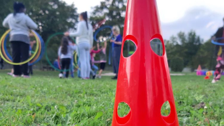 The picture shows a red cone standing on a lawn. Children playing with hula hoops can be seen in the background.