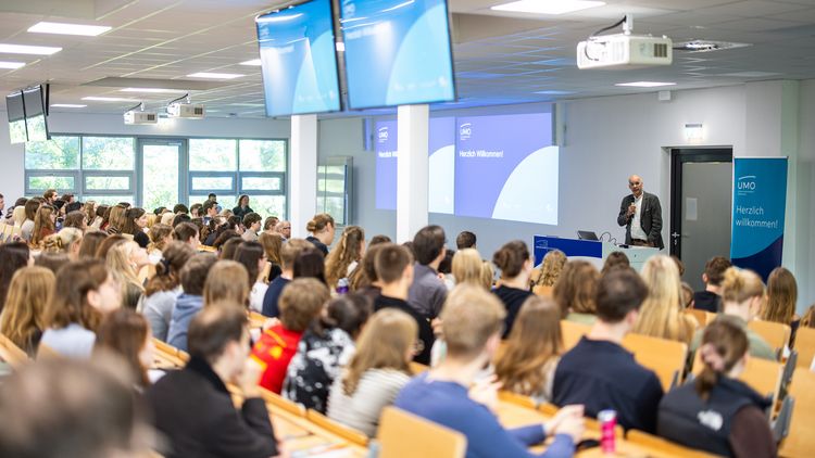 A well-filled lecture theatre. The students can be seen from the back. At the front stands a man with a microphone. Next to him is a roll-up banner bearing the words: UMO – A warm welcome.