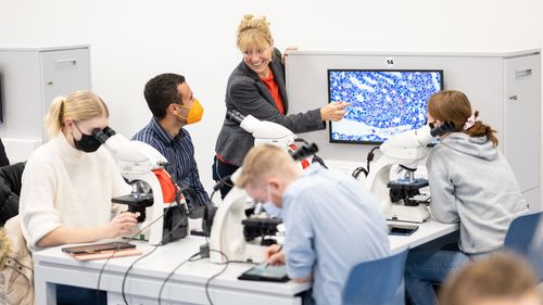 Four students sit at the group table at microscopes. Professor Anja Bräuer explains the image that is being transmitted from one of the microscopes to a monitor at the group table.