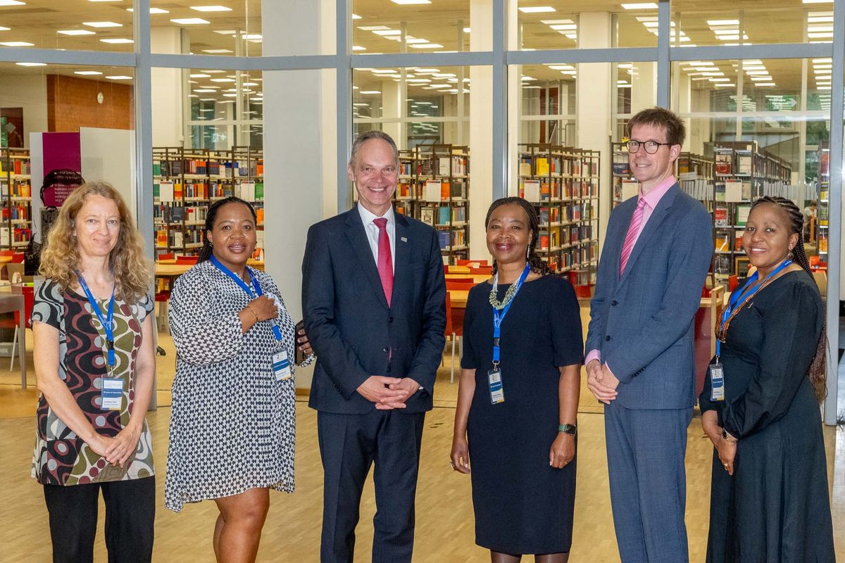  Das Bild zeigt eine Gruppe von sechs Personen, die vor der Bibliothek der UOL stehen und in die Kamera lächeln. Im Hintergrund sind hohe Bücherregale mit vielen Büchern zu sehen. Die Personen tragen formelle Kleidung und Namensschilder mit der Aufschrift "25 Years Partnership" und den Logos der Universitäten. Es handelt sich um Vertreter*innen der Universität Oldenburg und der Nelson Mandela University, die anlässlich des 25-jährigen Kooperationsjubiläums zusammengekommen sind. 
