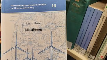 Ein etwas abgegriffenes Buch, auf dem blau-weißen Titelblatt sind Windräder zu sehen. Im Hintergrund Bücherregale der Bibliothek.