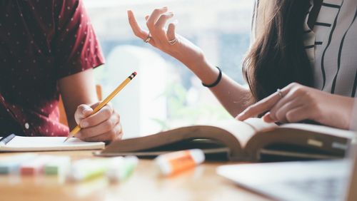 Zwei Jugendliche diskutieren miteinander über ein Buch, zu sehen sind nur die gestikulierenden Hände, das auf dem Tisch liegende Buch und eine weitere Hand, die einen Stift hält.