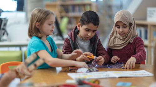 Three children of primary school age sit next to each other at a table and do handicrafts.