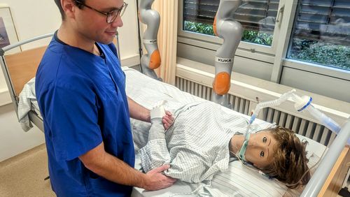Ein junger Mann im Klinik-Outfit steht an einem Krankenbett und hält die Hand der Roboterpatientin, die darin liegt.