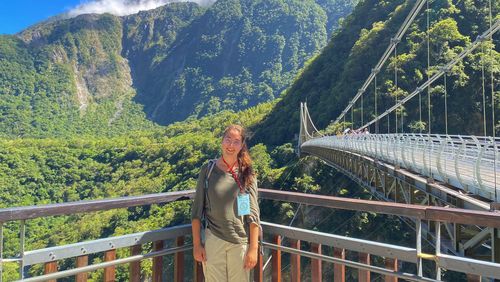 Selfie auf einer Aussichtsplattform vor bewaldeten Bergen, seitlich ist eine Hängebrücke zu sehen. 