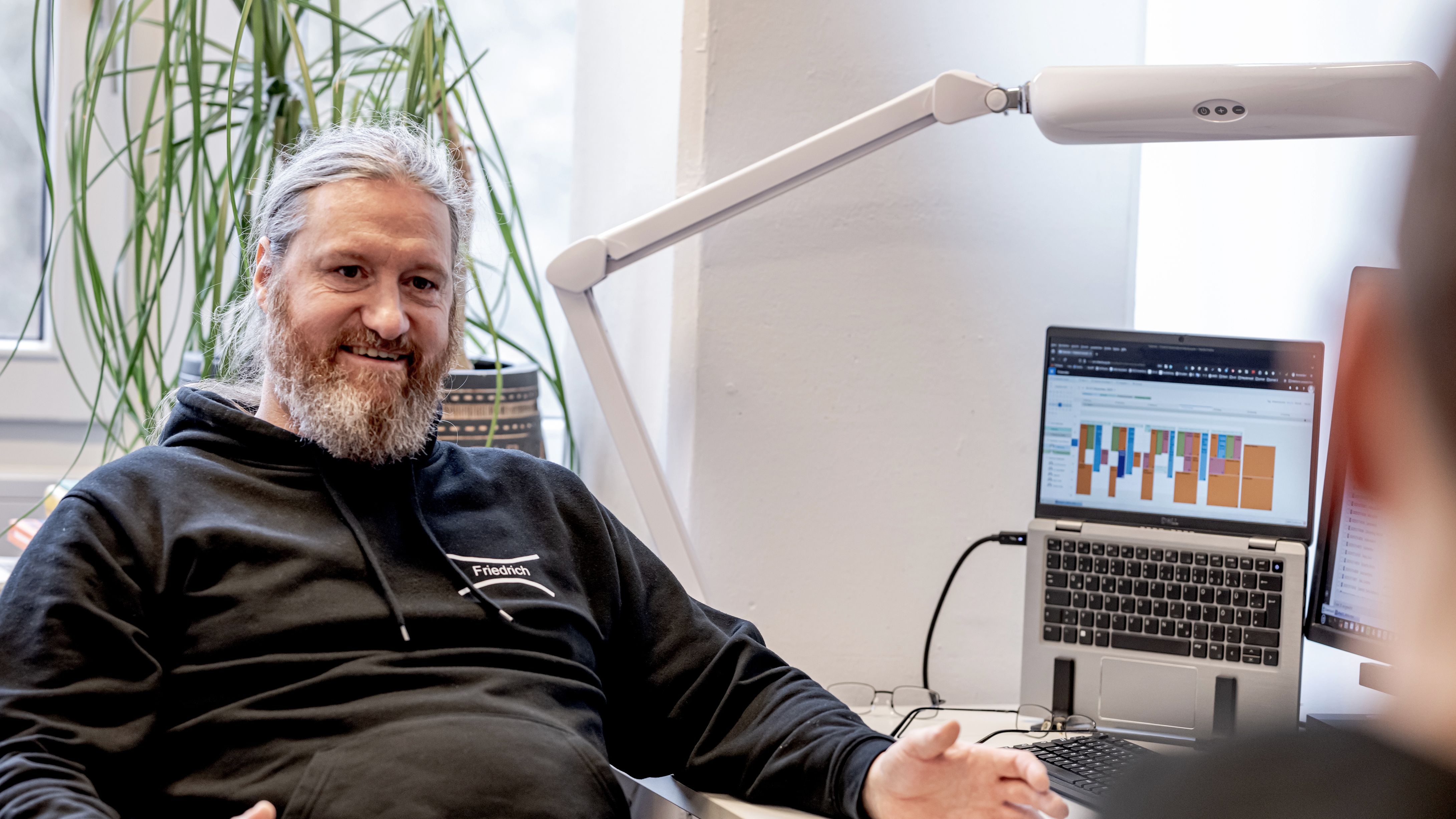 The picture shows Friedrich Hinrichs. He is sitting opposite the interviewer and explains something to him. He gestures slightly with his hands. To his right is an open laptop showing colourful rows, and behind him is a potted plant.