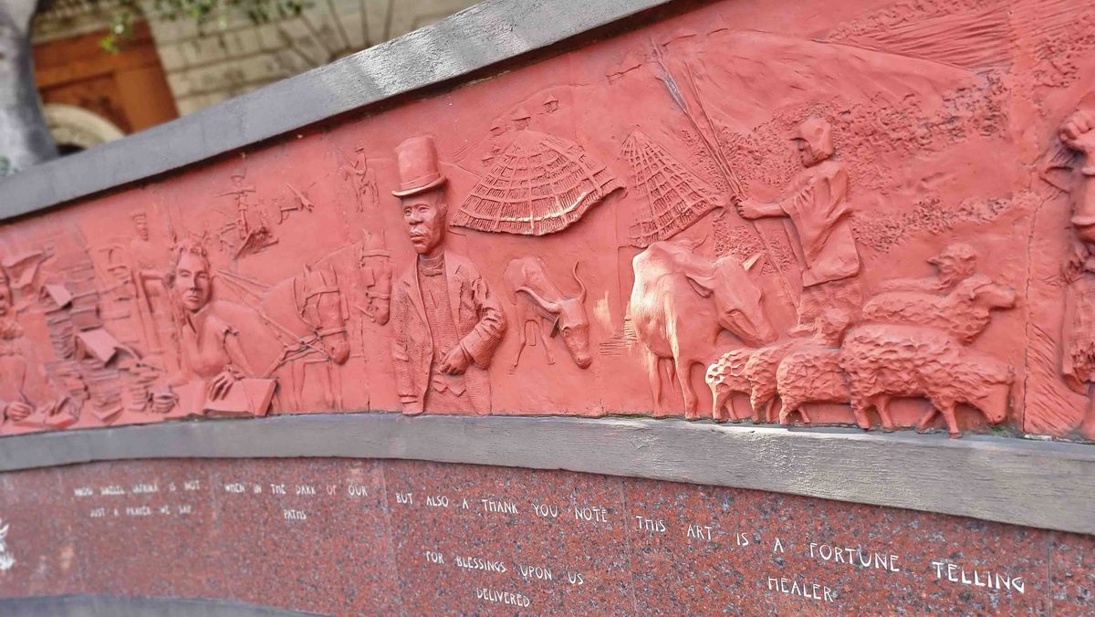 Ein rotes Relief an einer Wand mit Figuren und Szenen. Unterhalb des Reliefs ein eingravierter Text.