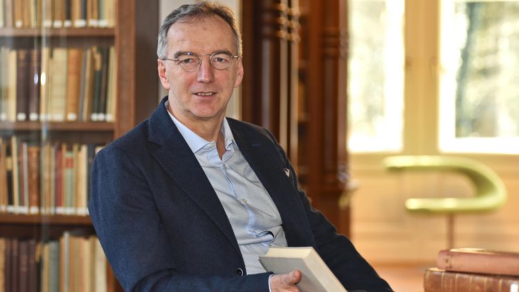 The picture shows Matthias Bormuth. He is sitting in the library of the Karl Jaspers House. Behind him stand shelves with books; he is holding a book in his right hand. Bormuth looks at the camera.