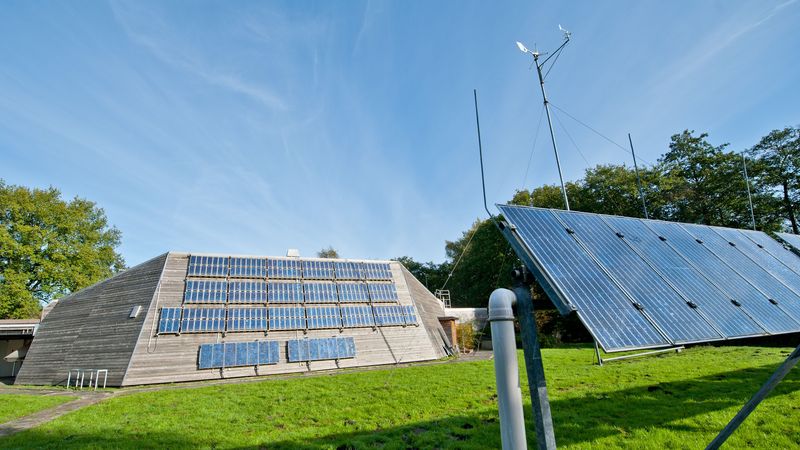 Außenaufnahme des Gebäudes, im Vordergrund Rasen und ein Gestell mit mehreren Solarmodulen, im HIntergrund Bäume.
