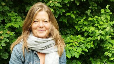 A woman with long, straight hair, a scarf around her neck and a denim jacket smiles at the camera.