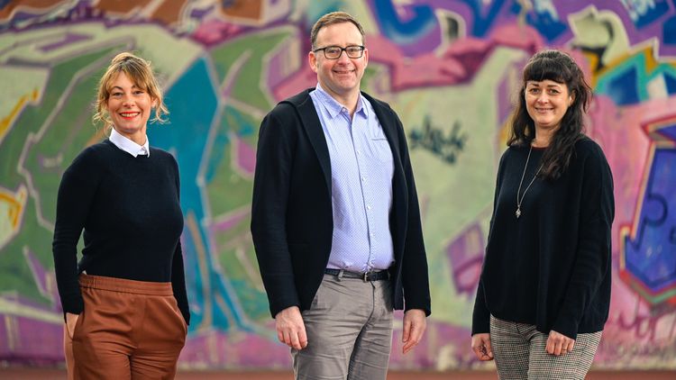 Three persons are standing in front of a wall covered with a graffiti.