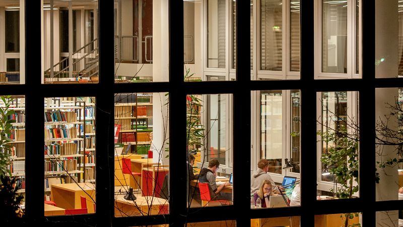 Das Bild zeigt die Bibliothek abends und von außen. In einem Raum kann man Menschen erkennen, die an Schreibtischen arbeiten. im Hintergrund stehen lange Regalreihen mit Büchern.