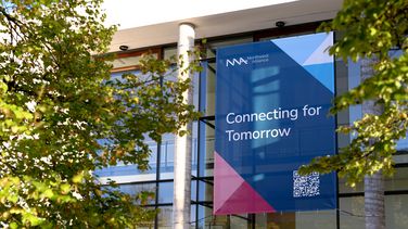 Großes Banner mit Text "Connecting for Tomorrow" hängt an Gebäudefassade des Hörsaalzentrums. Grüne Baumkronen ragen links und rechts ins Bild. 