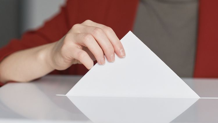 Nahaufnahme einer weiblichen Hand, die einen Stimmzettel in eine Wahlurne steckt.