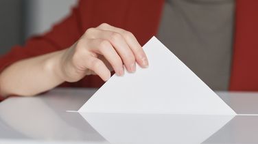 Nahaufnahme einer weiblichen Hand, die einen Stimmzettel in eine Wahlurne steckt.