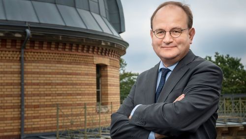 Das Bild zeigt Ottmar Edenhofer. Er steht mit verschränkten Armen draußen auf dem Dach eines Gebäudes. Hinter ihm ist ein runder Bau zu erkennen, der wie eine Sternenwarte aussieht. Edenhofer schaut in die Kamera.