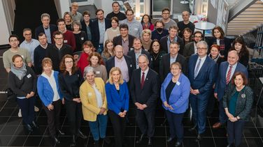 The picture shows the members of the senates and management teams of both universities. They are standing in a corridor of the HWK and smiling at the camera, which is looking down slightly on the scene.