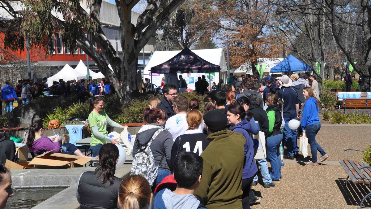 Eine belebte Veranstaltung auf dem Campus der University of New England. Menschen stehen in Schlangen vor Ständen oder sitzen. Im Hintergrund Zelte und Universitätsgebäude.