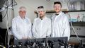 Three men are standing in a laboratory. All three are wearing white coats, a cap over their hair and special glasses to protect their eyes from the laser light. The test setup can be seen on a table in the foreground.