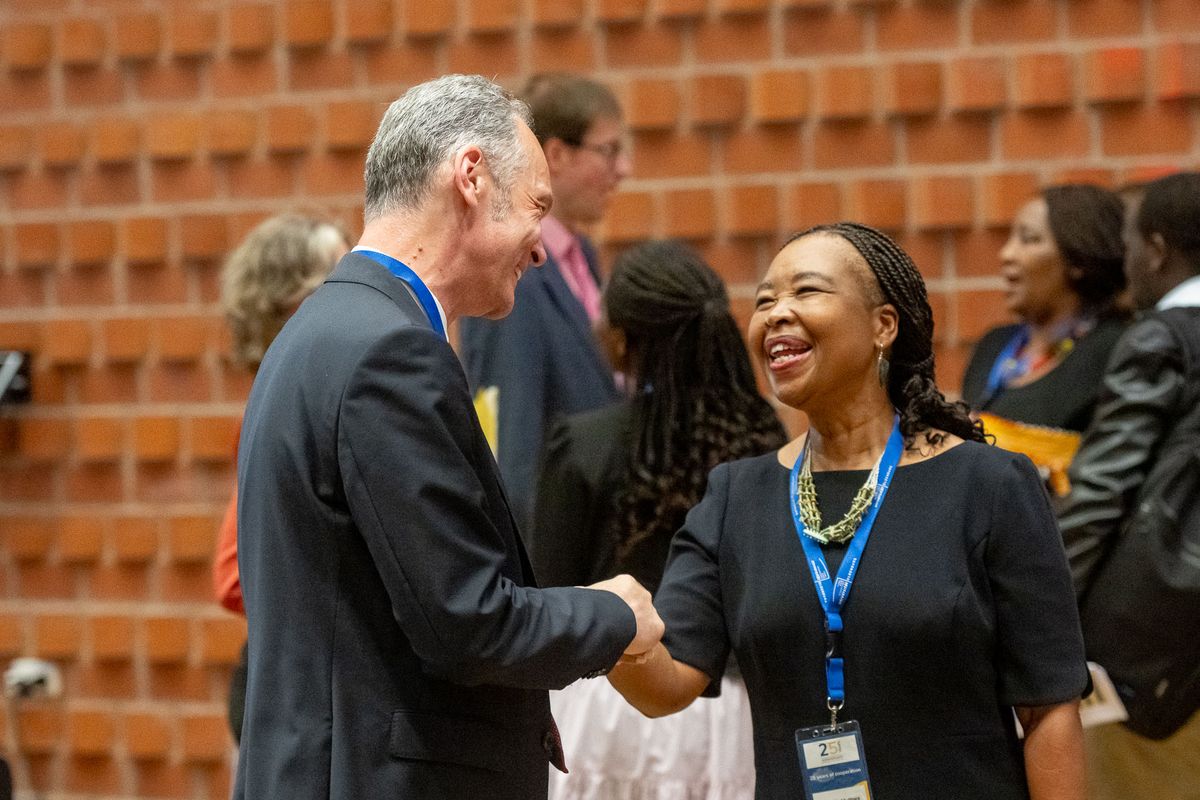 Das Bild zeigt zwei Personen, den Präsidenten der UOL und die Vice-Chancellor der NMU, die sich die Hände schütteln und lächeln. Beide lachen herzlich. Beide tragen Namensschilder mit der Aufschrift "25 Years Partnership". Im Hintergrund sind weitere Personen zu sehen, die sich ebenfalls unterhalten. Die Wand im Hintergrund besteht aus roten Ziegeln. 