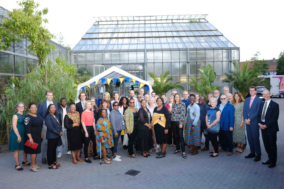 Das Bild zeigt eine große Gruppe von Menschen, die für ein Gruppenfoto posieren. Die Gruppe besteht aus Teilnehmenden des Welcome Dinners zum 25-jährigen Kooperationsjubiläum der Universität Oldenburg mit der Nelson Mandela University. Im Hintergrund ist ein großes Gewächshaus zu sehen, das mit Lichtern und bunten Fahnen dekoriert ist. 
