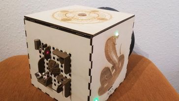 Edge view on a wooden cube. On one side, there is an image of a cobra, on the other a sun wheel, on the third plastic models of Aztec buildings. Not to be seen is the fourth side with a vault opening.